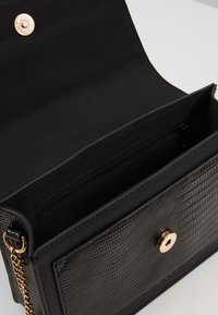 Czarna torebka o strukturalnym kształcie z teksturowanym wykończeniem, złotymi elementami, zapięciem na zatrzask oraz przestronnym wnętrzem, w tym kieszenią na zamek.