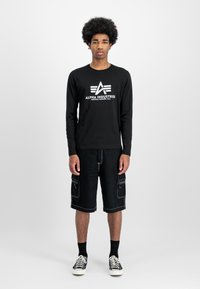 Svart langermet skjorte med hvitt logo, kombinert med svarte cargo-shorts med sidelommer, og svarte sokker med mønstrede sneakers.