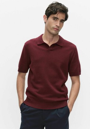 Jonge man met donker haar, gekleed in een bordeauxrode polo met korte mouwen en donkerblauwe broek, staand met handen in de zakken tegen een witte achtergrond.