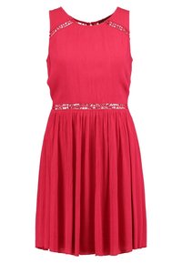 Robe rouge sans manches avec jupe plissée et bordure de sequins en forme de cœur au niveau du décolleté et de la taille, réalisée en tissu texturé.