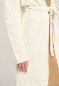 Cremefarbener gestrickter Cardigan mit einem Bindegürtel, der aus einem strukturierten Stoff und gerippten Bündchen besteht. Warmes, weiches Material mit einem gemütlichen Aussehen.