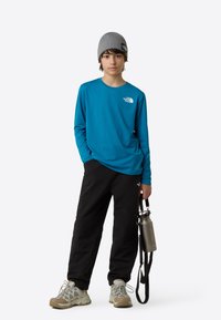 Modra majica z dolgimi rokavi z logotipom, črne hlače, bež pohodniški čevlji, siva kapa ter drži sivo steklenico za vodo z vrvico.