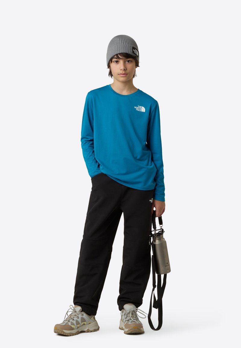 Modra majica z dolgimi rokavi z logotipom, črne hlače, bež pohodniški čevlji, siva kapa ter drži sivo steklenico za vodo z vrvico.