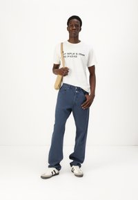 Witte T-shirt met zwarte tekst, blauwe denim jeans en beige schoudertas. Schoeisel met witte en zwarte accenten. Eenvoudig, casual ontwerp.