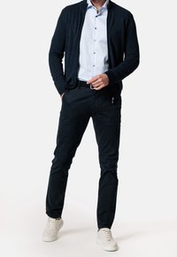 Tmavě modrý cardigan na zip přes světle modrou košili, doplněný tmavými kalhotami a bílými teniskami. Hladk�á látka, volný střih, minimalistický design.