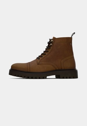 Brown leather lace-up ankle boot with thick black treaded sole and pull tab on a white background.