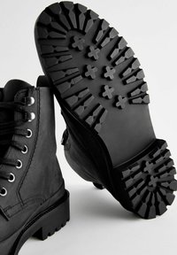 Botas de cuero negro con una suela de goma texturizada que presenta surcos profundos y patrones cruzados. Los cordones con ojales metálicos mejoran la durabilidad.