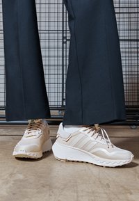 Beiga sportskor med tre randiga sidor, tyg- och gummidetaljer, texturerad sula, använda med mörka skräddarsydda byxor.