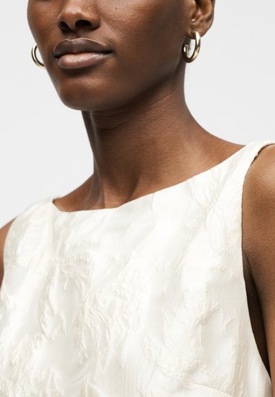Κοντινό πλάνο γυναίκας που φορά χρυσά σκουλαρίκια κρίκους και λευκό αμάνικο τοπ με υφή και λαιμόκοψη σκάφος.