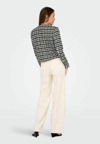 Melnbalts houndstooth raksts žakete, īsā designā, valkāta ar krēmkrāsas platām biksēm un melniem zābakiem ar papēžiem, izceļot gludu tekstūru.