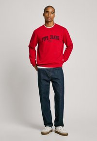 Sudadera roja con el logo "PEPE JEANS" en azul marino, combinada con jeans de color azul oscuro y zapatillas blancas. Diseño de corte relajado y cuello redondo.