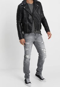 Chaqueta de motorista de cuero sintético negra con cremallera plateada, camiseta gris, jeans gris desgastados y zapatillas altas negras con suelas blancas.