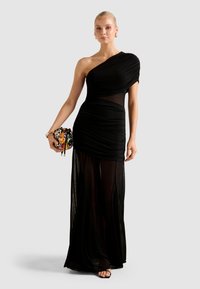 Sieviete tērpusies vienā plecā melnā caurspīdīgā kleitā, rokās turot mazu, krāsainu fliteru somiņu, stāv pret vienkrāsainu gaiši pelēku fonu.