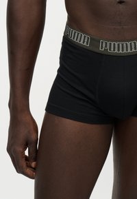 Torso et parties supérieures des jambes d’un homme portant un boxer Puma noir avec une ceinture verte et le logo Puma, sur un fond clair uni.