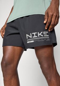Melnas sporta šortas ar acu paneļiem, kas rotāti ar lielu pelēku Nike logo un tekstu kreisajā apakšējā pusē. Viegls audums un īsa griezuma modelis.