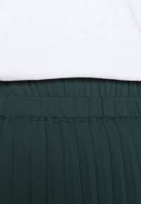 Grøn plisseret stoflinning med folder, under en hvid, blød t-shirt kant. Ingen tydelige mønstre eller udsmykninger synlige.