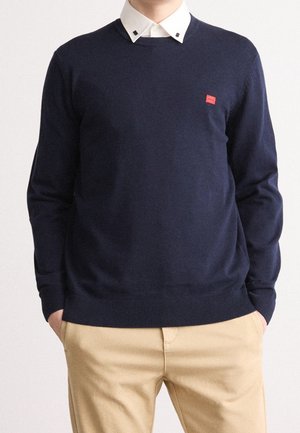 Uomo che indossa un maglione blu navy sopra una camicia bianca con colletto e pantaloni beige, con le mani nelle tasche, in piedi davanti a uno sfondo chiaro e semplice.