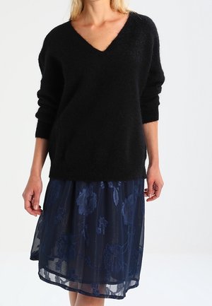 Schwarzer, flauschiger V-Ausschnitt-Pullover kombiniert mit einem dunkelblauen, durchsichtigen Rock mit floralen Stickereien. Lange Ärmel und lässige Passform.