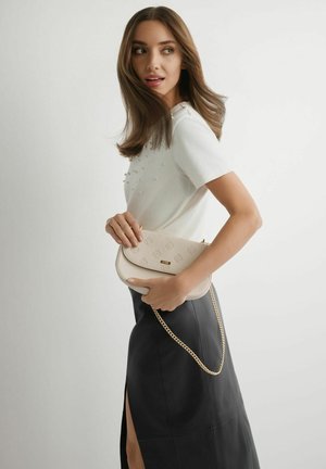 Kobieta ubrana w biały top z krótkim rękawem i czarną spódnicę z rozcięciem z boku, trzymająca beżową torebkę z złotym łańcuszkiem na jednolitym tle.