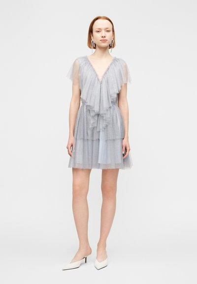 Γυναίκα με κοντά κόκκινα μαλλιά, ντυμένη με ένα ανοιχτό μπλε, λαμπερό, πολυεπίπεδο φόρεμα από τούλι και λευκές μυτερές γόβες, στέκεται μπροστά σε ένα λευκό φόντο.