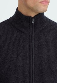 Zwart zip-up sweater met een hoge kraag, textuur op het oppervlak en een verticale ribbelpatroon. Voorzien van een voorrits voor sluiting.