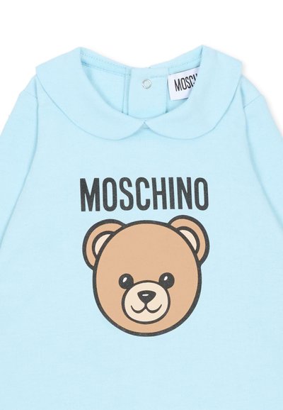 Camicia in tessuto azzurro chiaro con una grafica di un orso marrone in stile cartone animato e la scritta "MOSCHINO" in stampa nera in grassetto. Include un colletto alla Peter Pan.