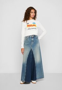Vrouw met krullend haar in een witte lange mouwen top met logo's en een twee-kleurige hoog-uiw denim maxi rok die staat met een hand op de heup.