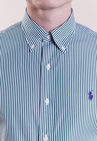 Grön-och-vitrandig skjorta med knappar, har en spridd krage, vita knappar och en liten lila logotyp på vänster bröst.