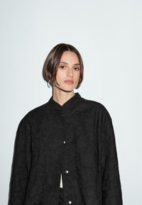 Kvinna med kort brunt hår och hoopsörhängen, iklädd en svart blommig texturerad jacka mot en enkel ljus bakgrund.