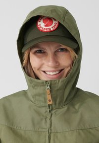 Olivgrön jacka med högt krage och dragkedja, med ett beige dragkedjehandtag. Personen bär en matchande grön keps med en rund logotyp.
