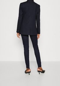 Tmavě modrý přizpůsobený blejzr s dlouhými rukávy, se strukturovaným tvarem a manžetami na jeden knoflík. V kombinaci s úzkými tmavými džíny a černými podpatky.