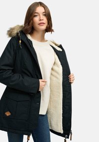 Svart parkajacka med huva i päls, dragkedjestängning och krämfärgad fuskpälsfoder. Har två framfickor och detaljer i brunt läder.
