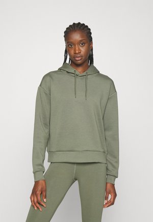 Femme aux cheveux tressés portant un sweat à capuche vert olive et des leggings assortis, debout devant un fond clair uni.
