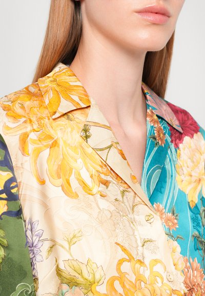 Woman wearing a colorful floral patterned blouse with yellow, blue, red, and green sections, featuring a pointed collar and button closure.