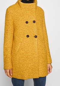 Abrigo amarillo de mezcla de lana con cuello alto, cierre cruzado con dos grandes botones y bolsillos laterales. La textura es suave y gruesa.