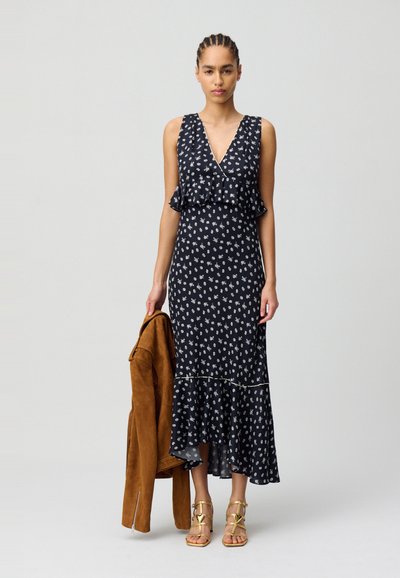 Vestido maxi floral en navy con acentos de volantes, que presenta un escote en V profundo y un dobladillo acampanado. La modelo sostiene una chaqueta de ante marrón y lleva sandalias doradas.