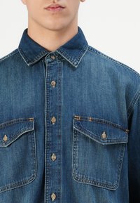 Homme portant une chemise en denim bleu à boutons avec deux poches poitrine et des coutures beige, vue de face recadrée au niveau de la bouche et des épaules.