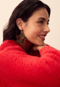 Green and red star-shaped earrings made of tinsel; worn with a textured red fuzzy sweater, showcasing intricate details and vibrant colors.