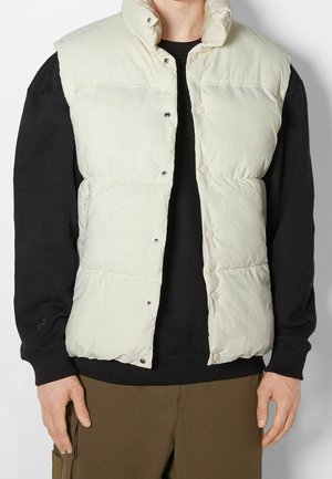 Personne portant une veste matelassée sans manches beige clair sur une chemise noire à manches longues et un pantalon marron, debout devant un fond blanc.