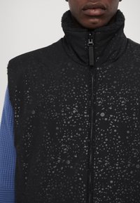 Black vest with a textured pattern of subtle droplets, zip closure, and a plush collar. Sleeves in contrasting blue knitted fabric.