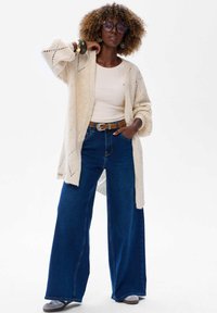 Crème breipully met ruitpatroon, beige aansluitende top, hoog-waisted wijde blauwe jeans en grijze instappers.