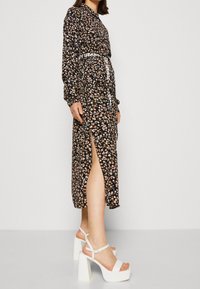 Robe noire à fleurs avec des manches longues, fente sur le côté et une ceinture avec "MY JEANS" en blanc. Portée avec des sandales à talons blocs blanches.