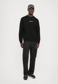 Svart genser med logo-patch, kombinert med svarte cargo-bukser med flere lommer. Grå caps og svarte sneakers fullfører antrekket.