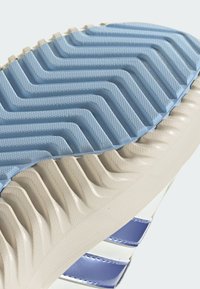 Potplat sportske tenisice s teksturiranim plavim slojem gume s valovitim uzorkom, koji prelazi u glatki kremasti donji sloj.