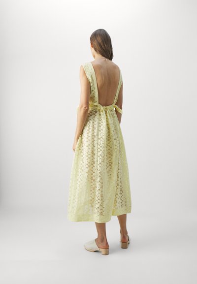 Robe dos nu jaune clair avec de larges bretelles, présentant un motif texturé et un détail de taille nouée. Associée à des chaussures à talons bloc neutres.