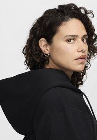 Μαύρο φούτερ με μεγάλο κουκούλι και κορδόνια. Το μοντέλο έχει σγουρά μαλλιά και μικρούς ασημένιους κρίκους στα σκουλαρίκια, με απλό και καθαρό φόντο.