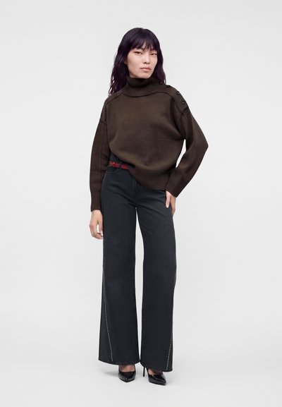 Femme portant un pull à col roulé marron foncé avec des boutons sur l'épaule, un pantalon noir large et des talons noirs, debout, main sur la hanche.