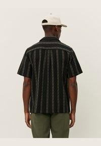 Black short-sleeve shirt with vertical white stripes, collared design, and textured fabric, paired with olive green pants and a light cap.