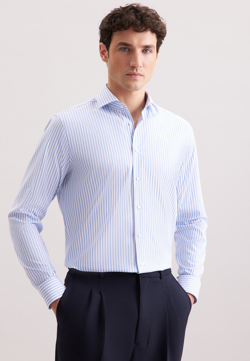 Homme portant une chemise habillée rayée bleu clair et blanche, rentrée dans un pantalon plissé noir, se tenant les mains dans les poches devant un fond uni.