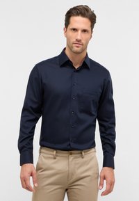 Navyblauw overhemd met lange mouwen en een button-down kraag, subtiele textuur, voorzak en zwarte knopen. G gedragen met lichtbeige broek.